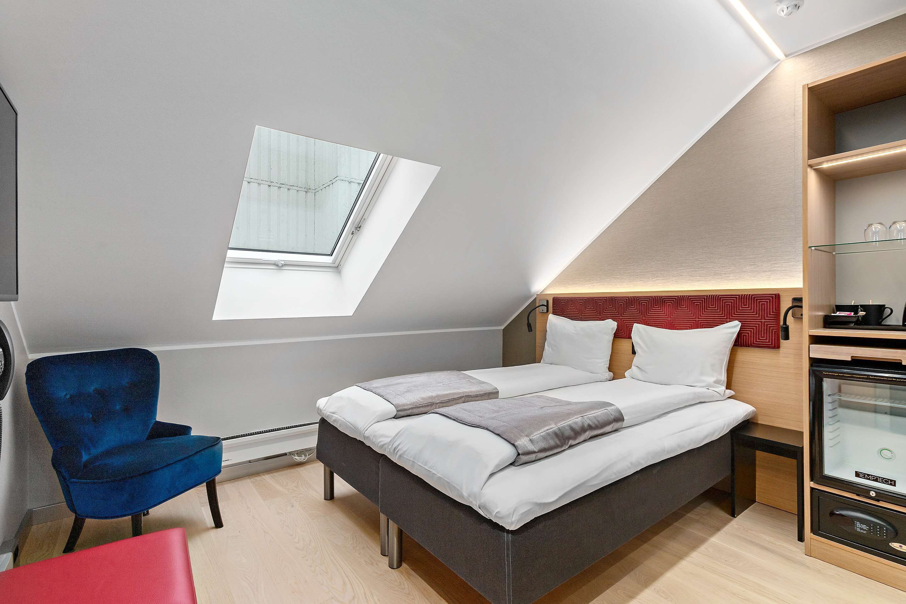 A modern bedroom with a skylight, a bed with white linens, a blue armchair, and a red ottoman.