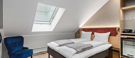 Un dormitorio moderno con tragaluces, una cama con ropa blanca, un sillón azul y un reposapiés rojo.