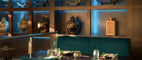 Un área de comedor con una mesa dispuesta para dos personas, un asiento tipo cabina tapizado en color verde azulado y adornos en estanterías detrás del asiento.