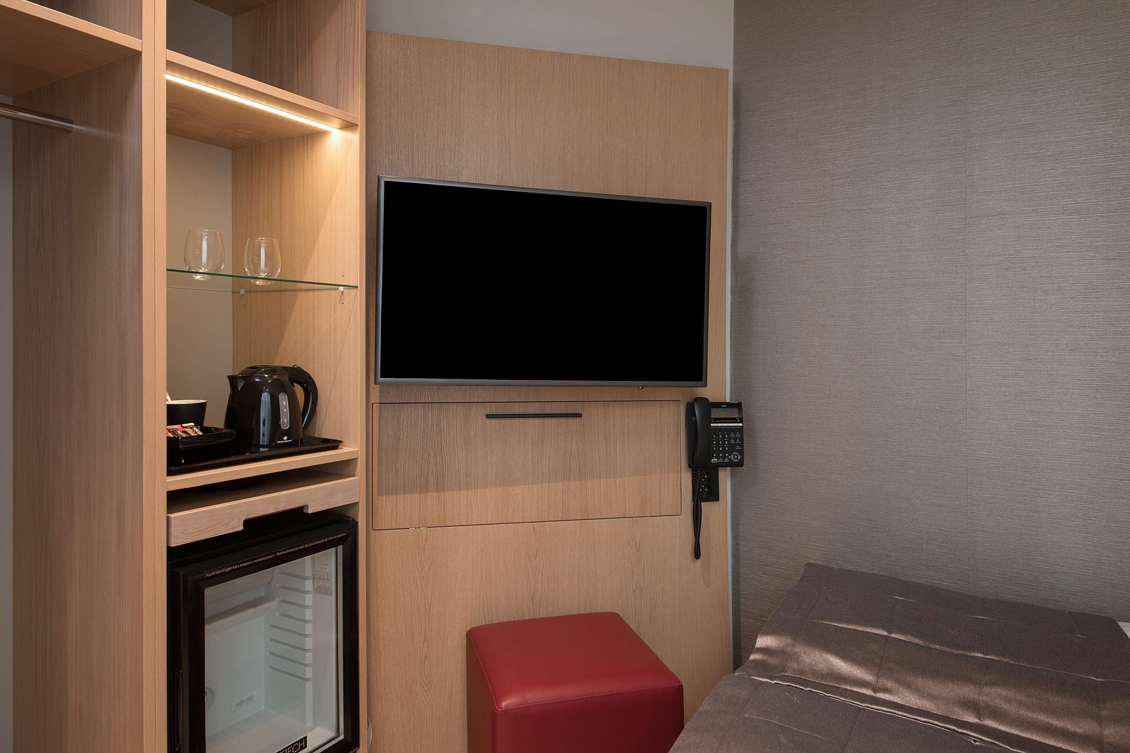 A compact room with a flat-screen TV mounted on the wall, a small refrigerator, a glass shelf with a kettle, and a red ottoman.