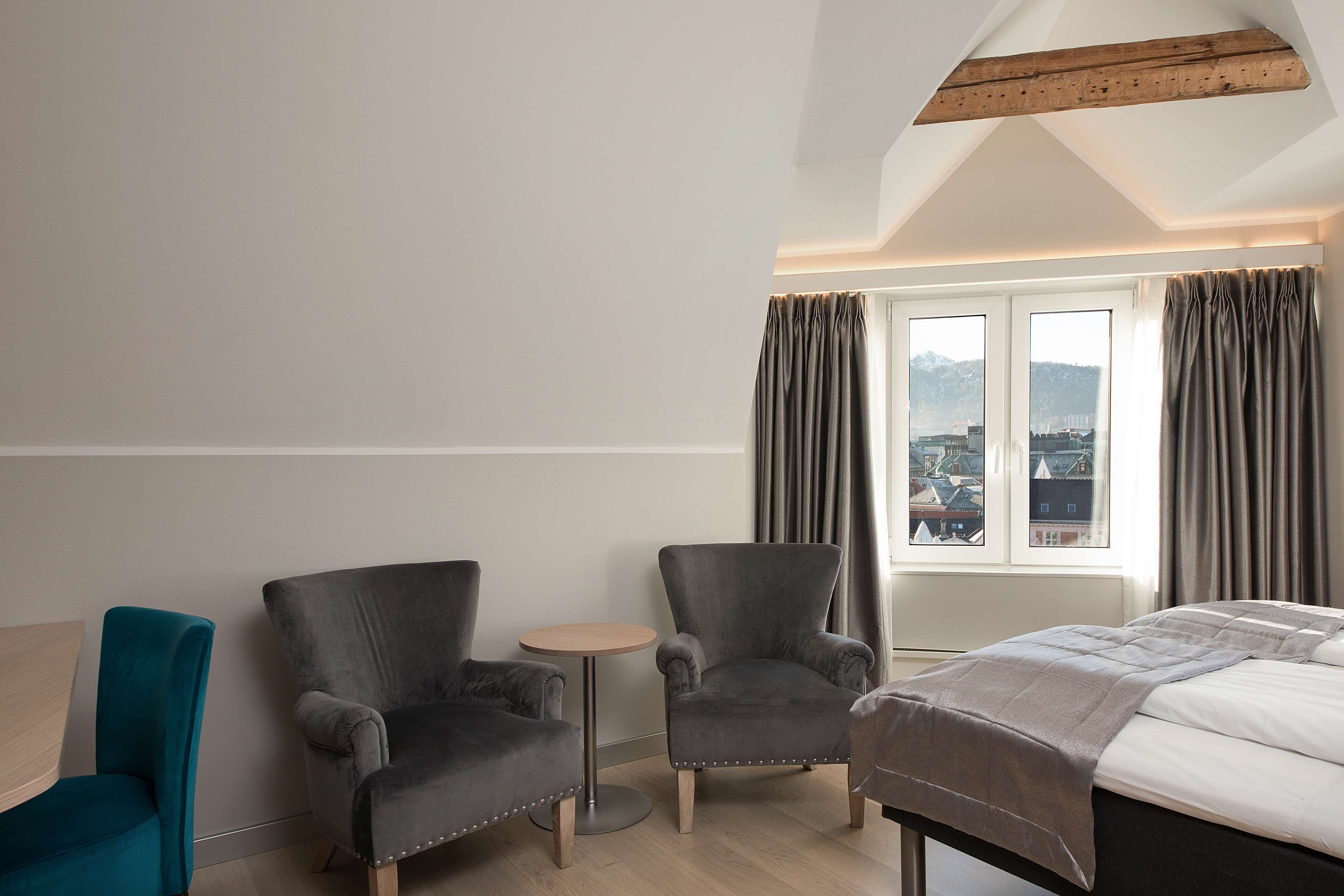 Une chambre d’hôtel avec un lit, deux fauteuils, une petite table ronde et une vue sur le paysage urbain, visible à travers de grandes fenêtres.