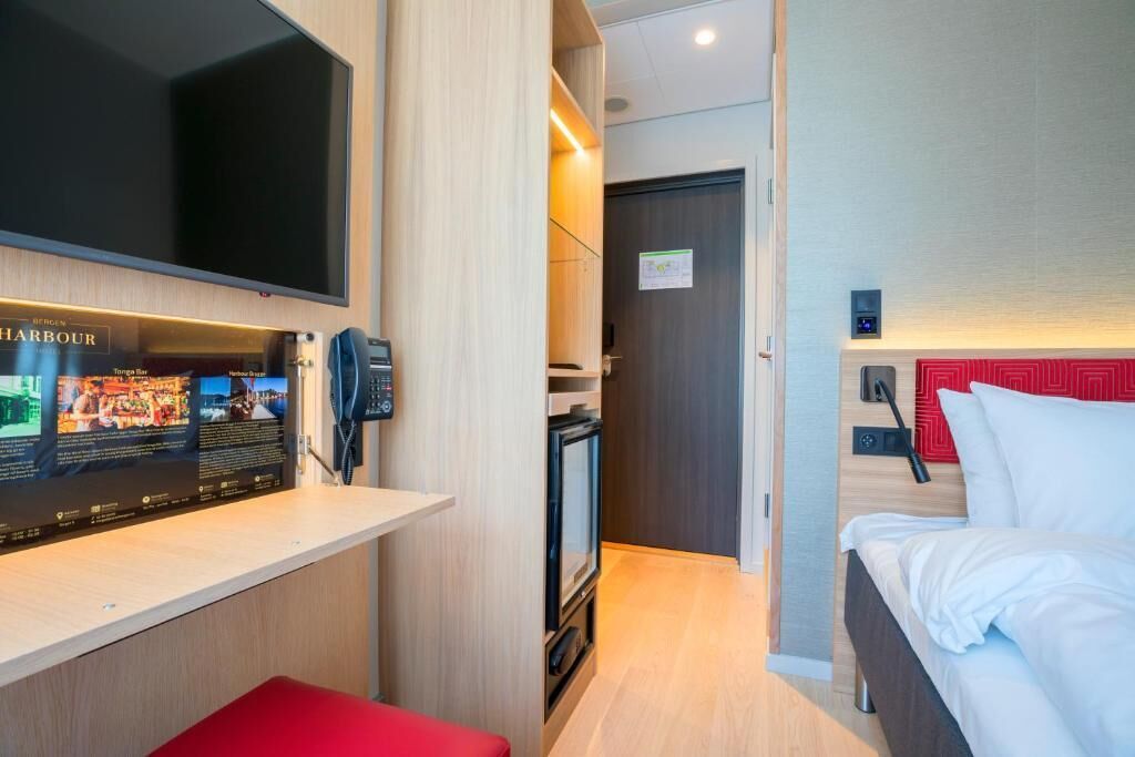 A modern hotel room with a flat-screen TV, a small kitchenette, and a bed with white bedding.