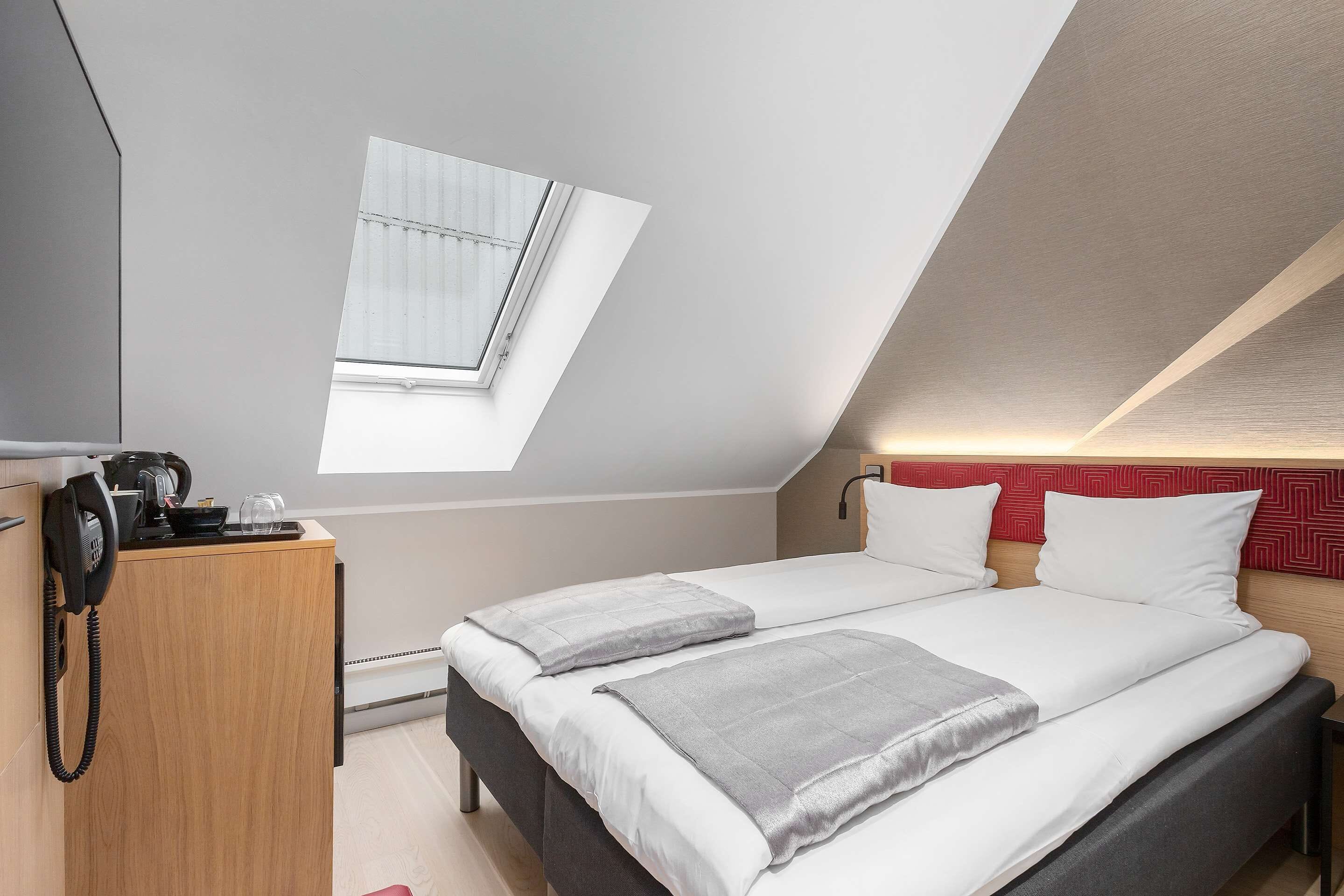 A hotel room with a bed, a desk with a telephone, and a skylight.