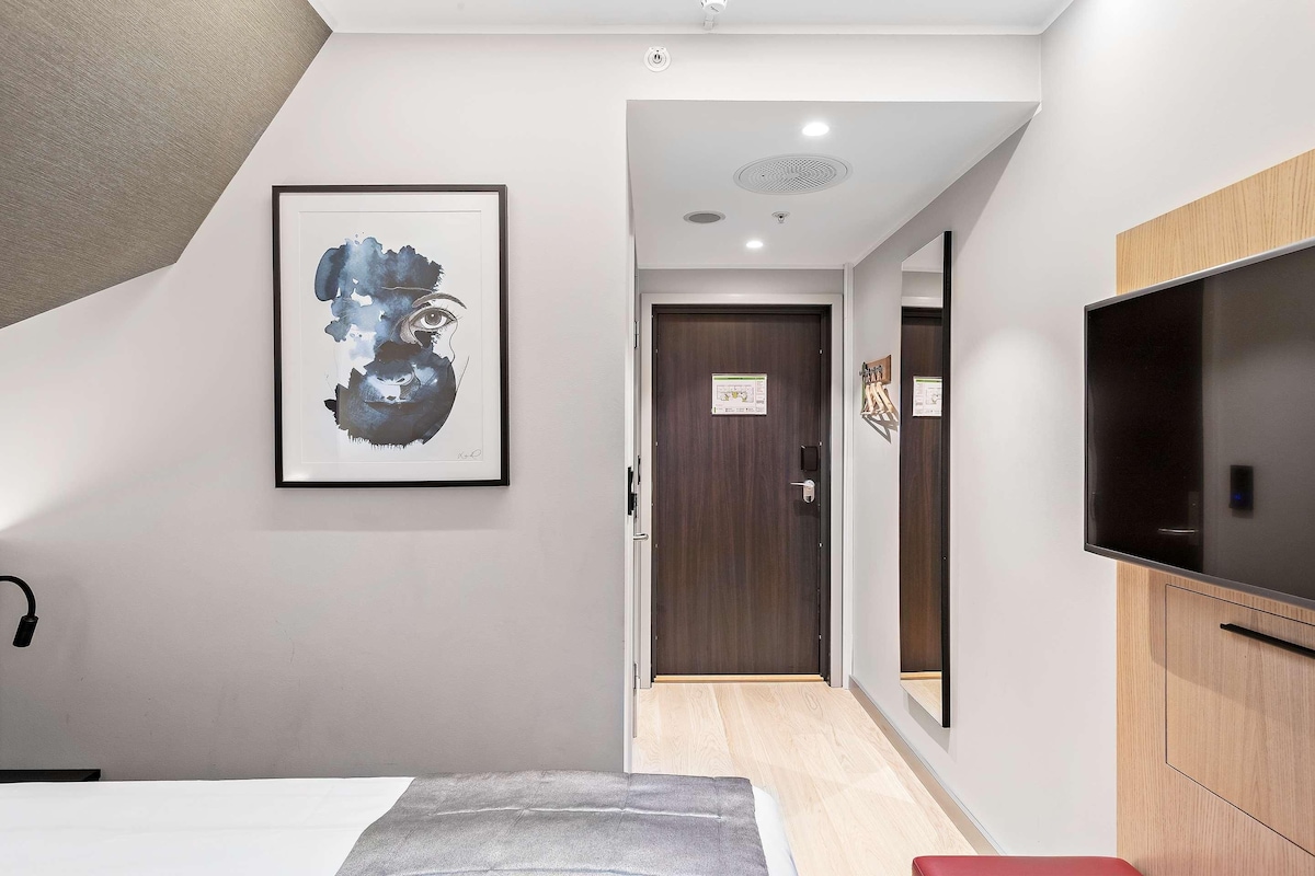A hotel room with a bed, a TV, a framed artwork, a wooden cabinet, and a door leading to another room.