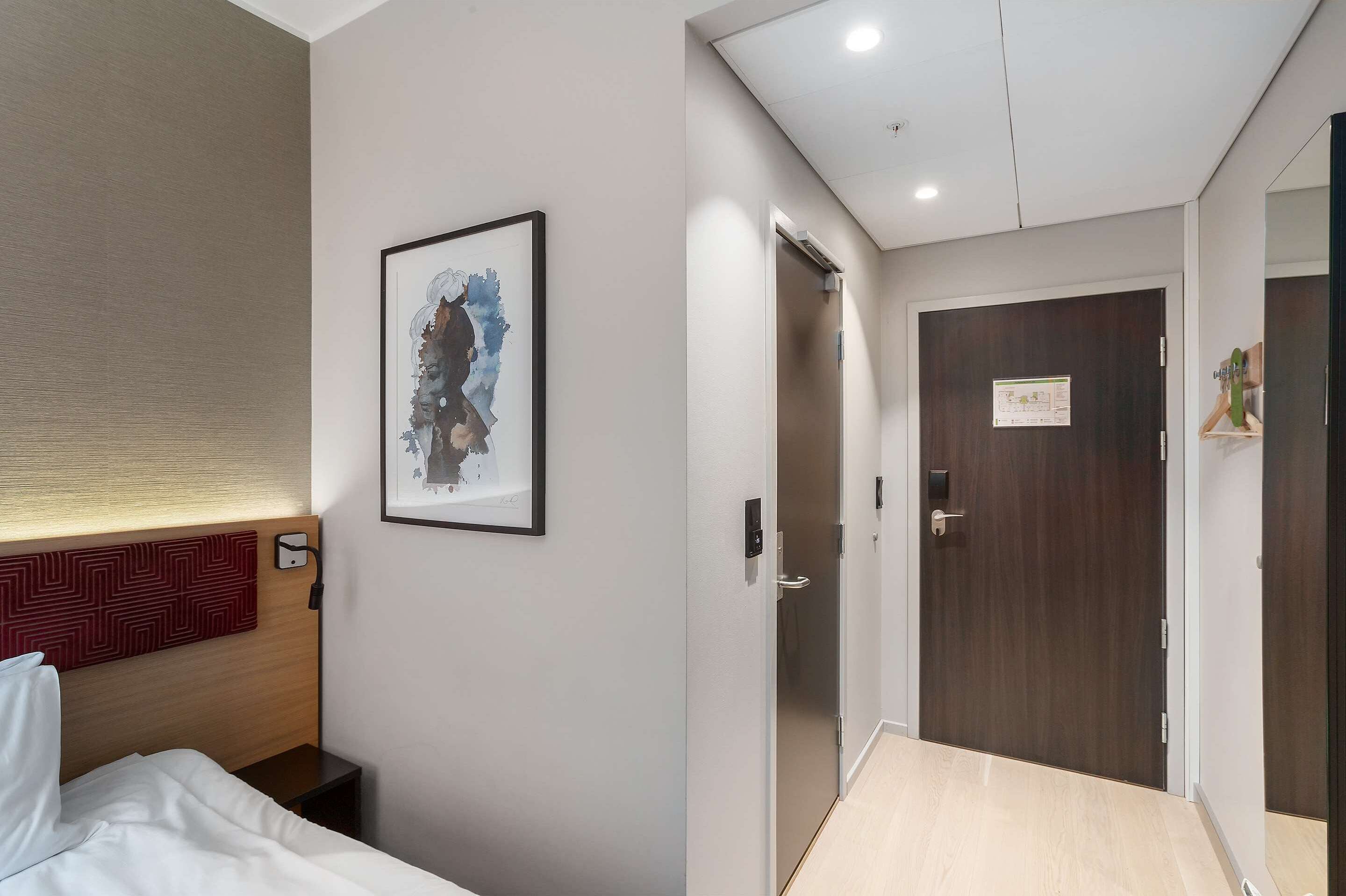 A hotel room with a bed, a headboard, a framed artwork, a wooden door, and a light switch.
