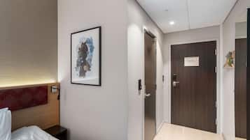 A hotel room with a bed, a headboard, a framed artwork, a wooden door, and a light switch.