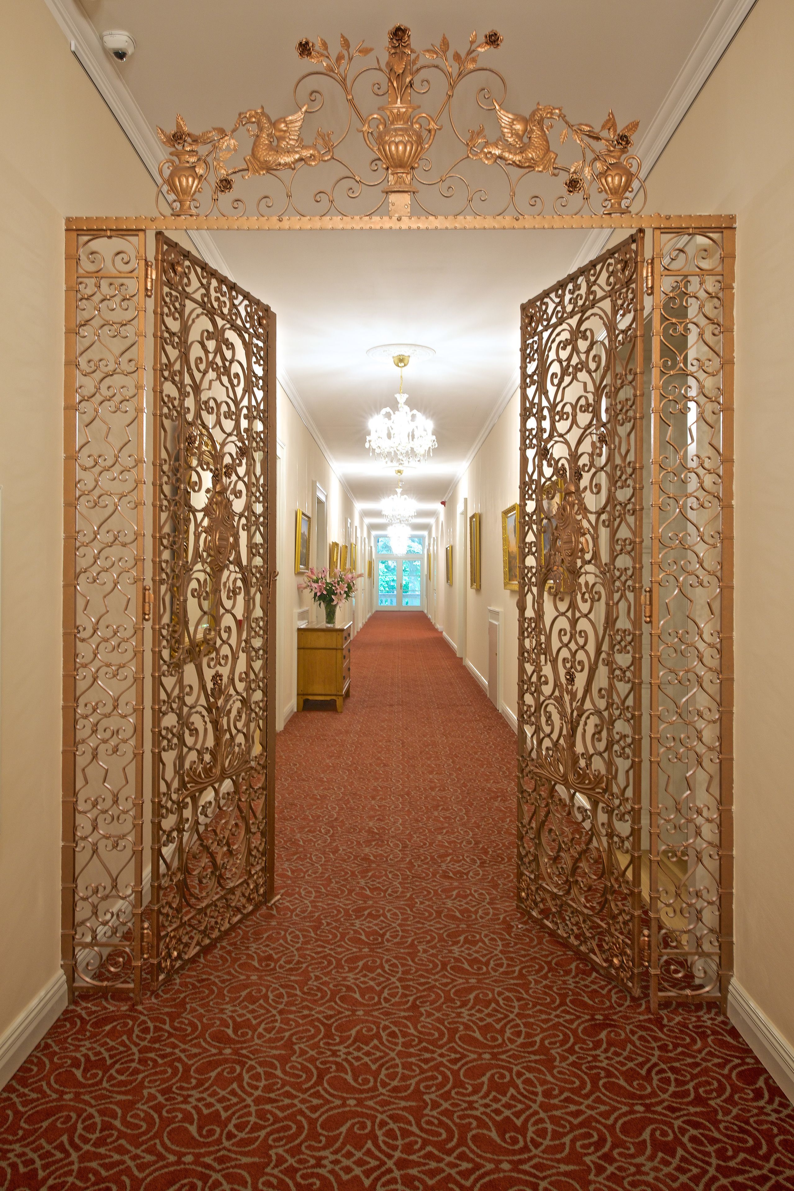 Un pasillo con puertas doradas ornamentadas, una alfombra roja con estampado y un candelabro.