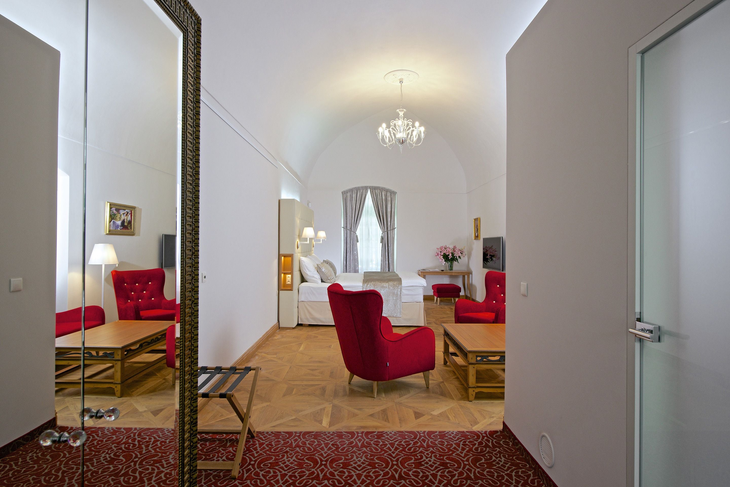 Een moderne hotelkamer met een grote spiegel, rode fauteuils, een houten eettafel en een kroonluchter.