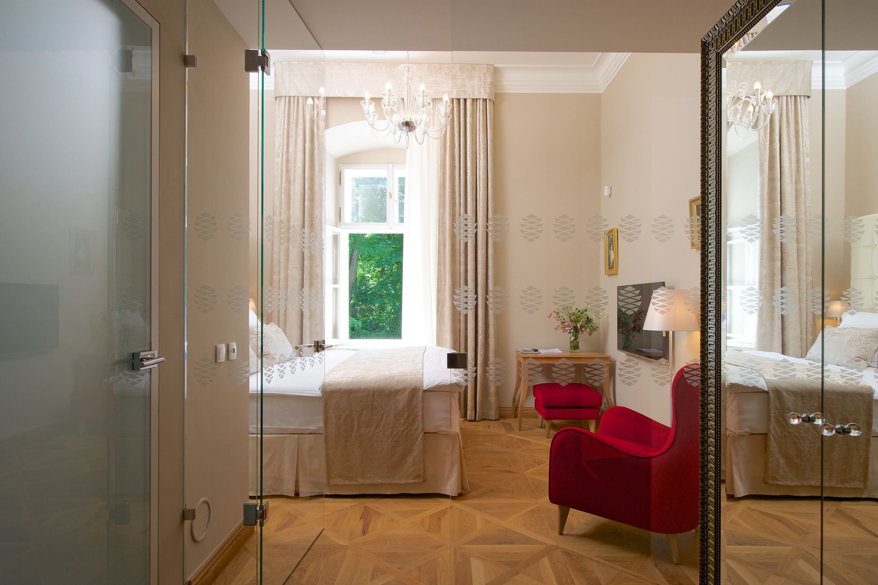 A hotel room with a bed, a red chair, a small table with a lamp, and a large mirror.