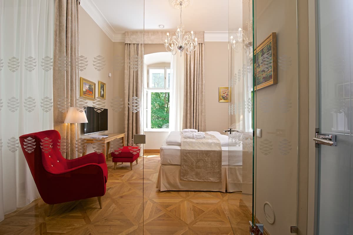 A hotel room with a bed, a red armchair, a TV, and a window with curtains.