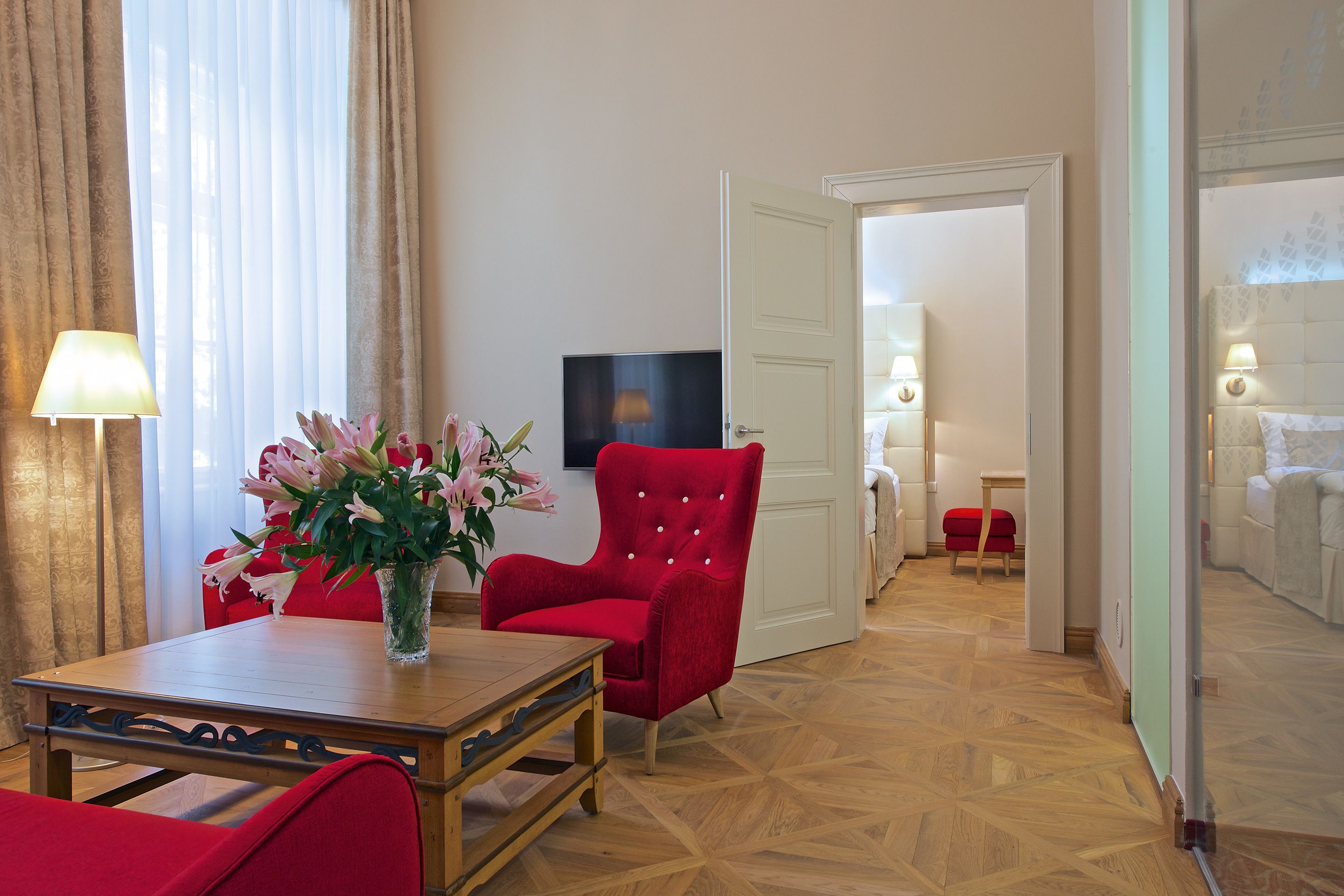 A hotel room with a wooden floor, a red armchair, a table with a vase of flowers, a television, and a bed with a white headboard.