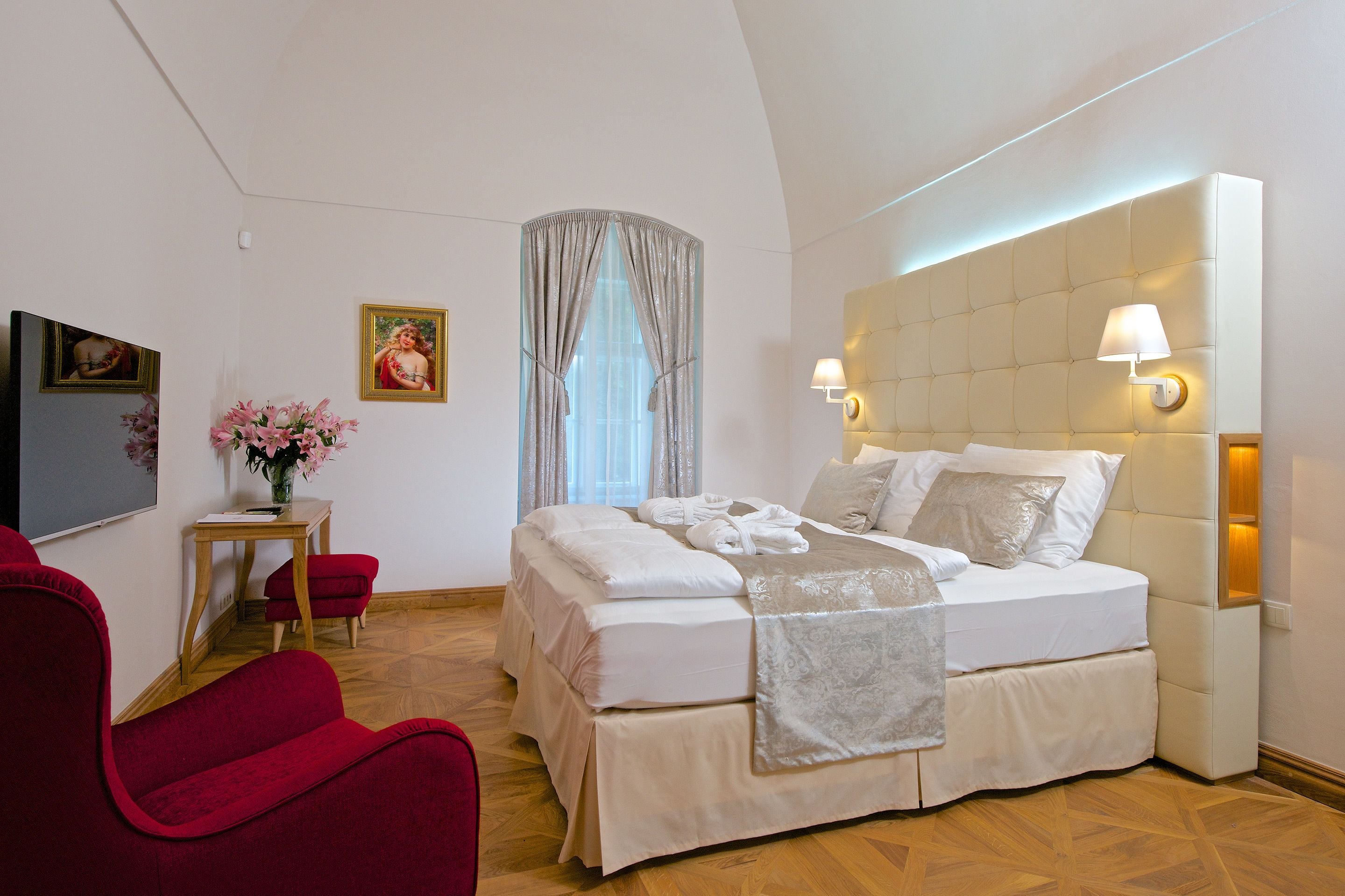 A hotel room with a large bed, a red armchair, a small table with a vase of flowers, and a painting on the wall.