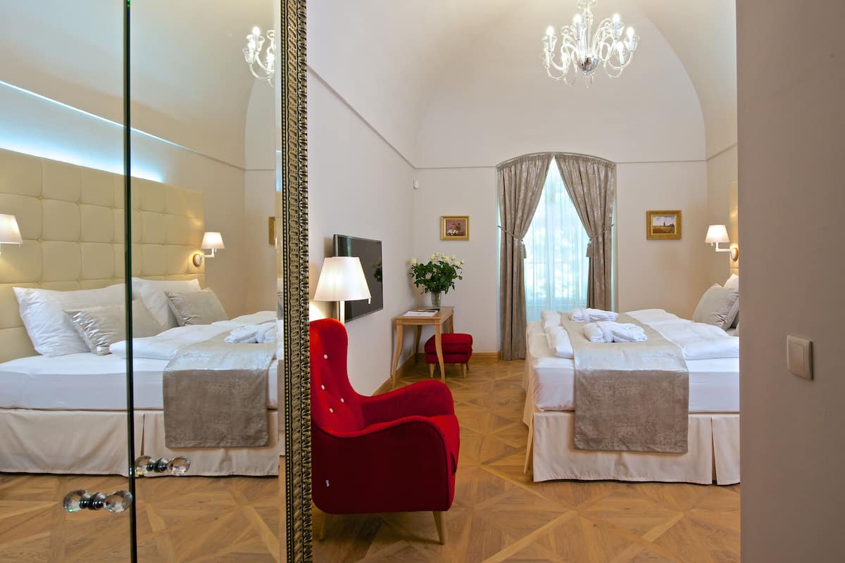A hotel room with two beds, a red chair, a TV, and a chandelier.