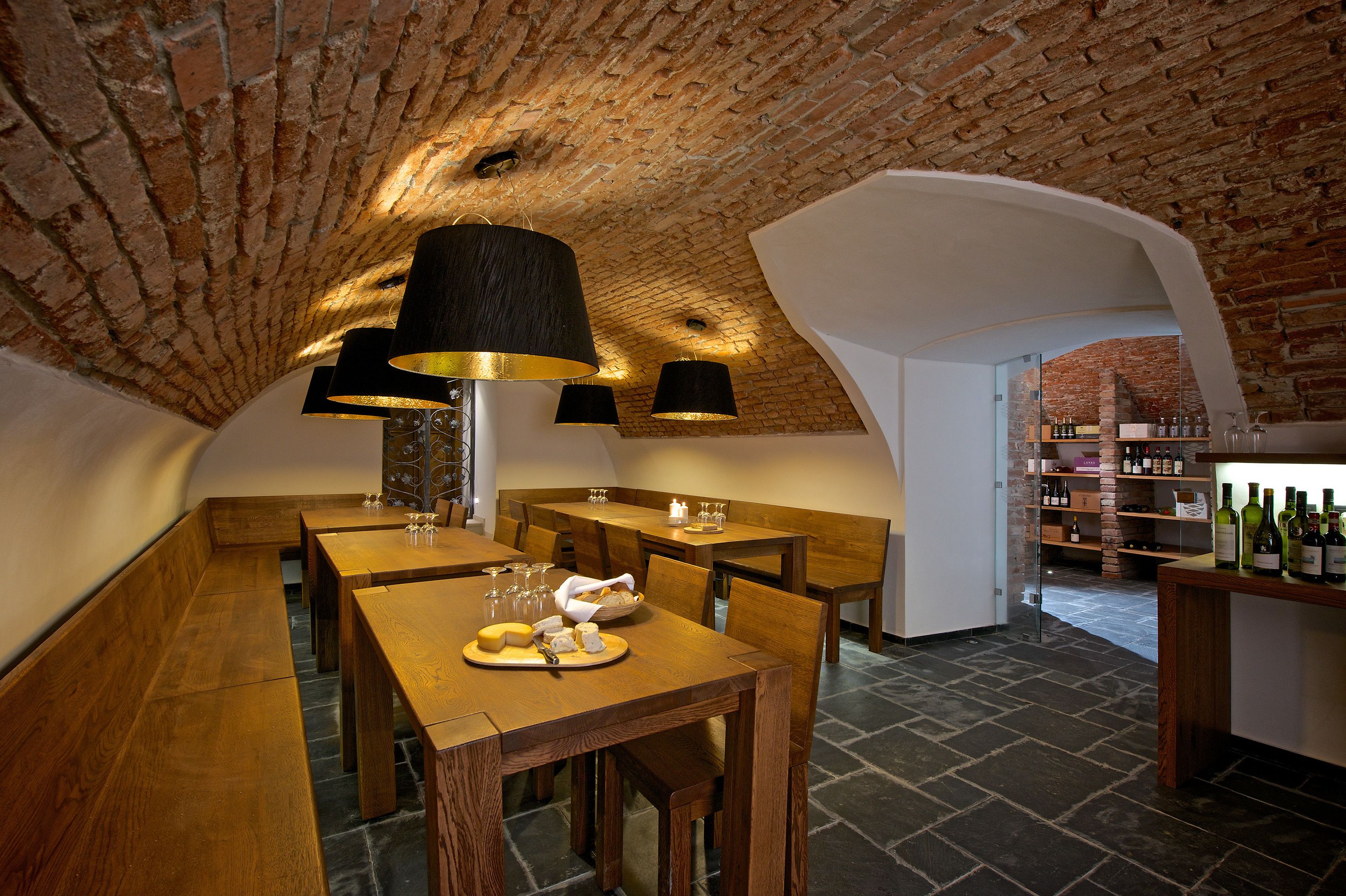 A rustic cellar with wooden tables and benches, a brick ceiling, and a wine rack with bottles.