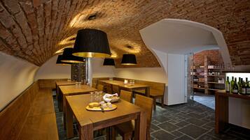A rustic cellar with wooden tables and benches, a brick ceiling, and a wine rack with bottles.