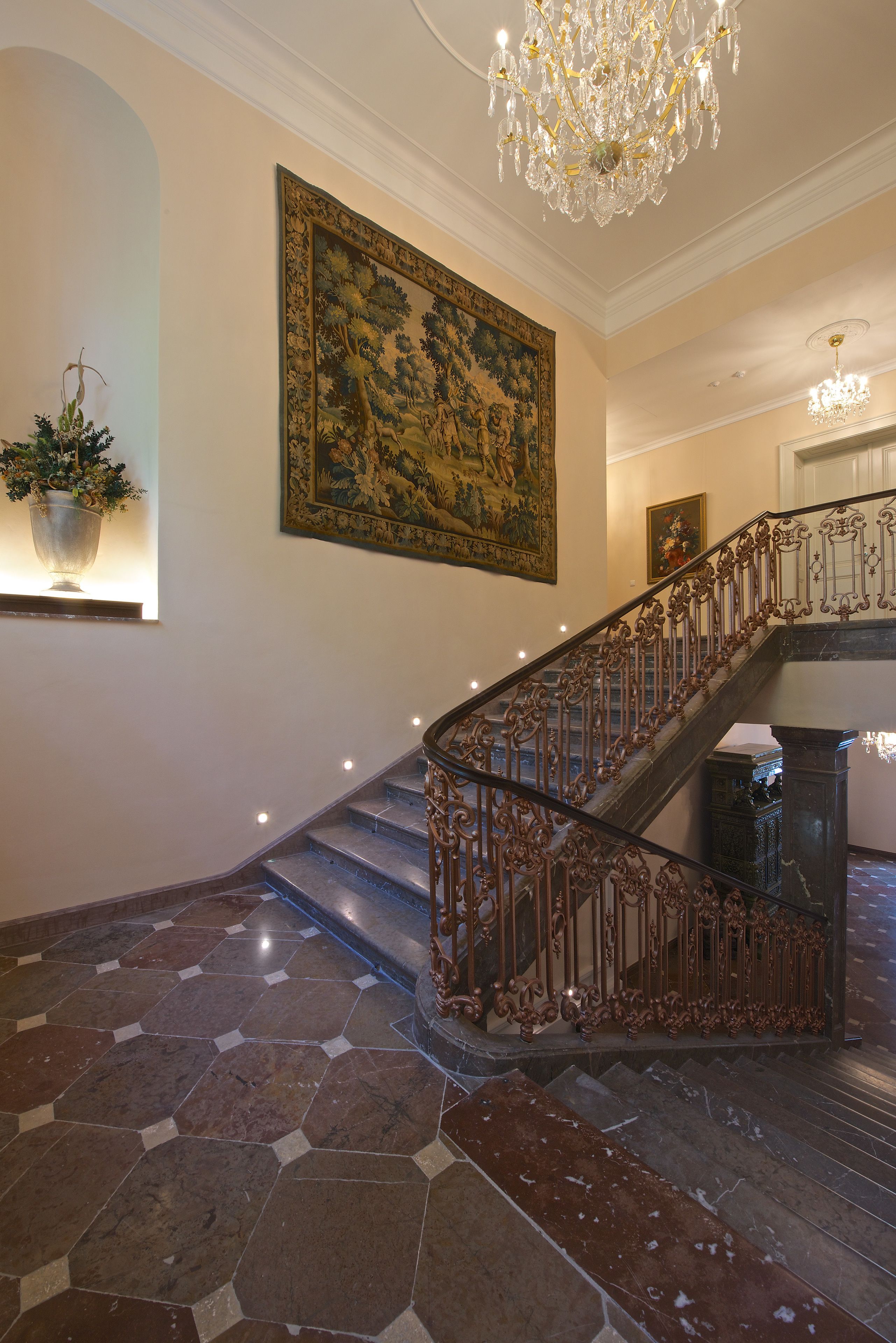 A grand staircase with ornate metal railings and a large, detailed painting on the wall.