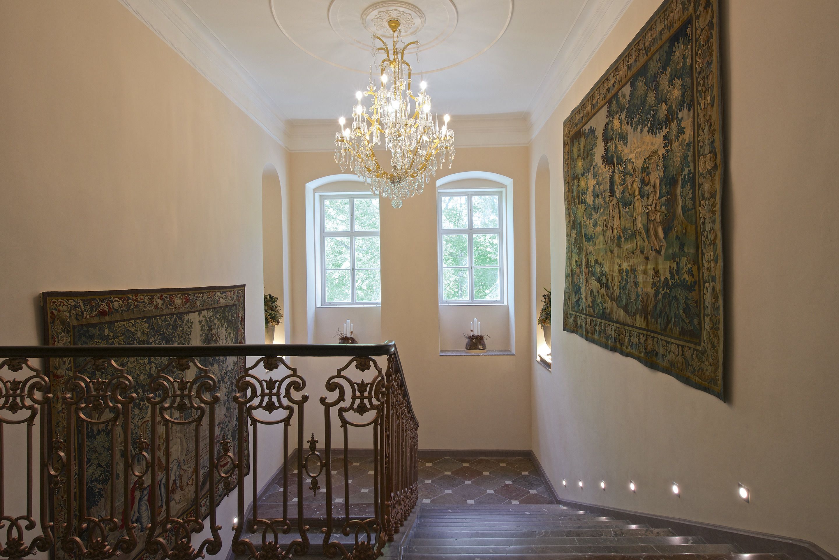 A grand staircase with ornate railings, a chandelier, and a large tapestry on the wall.