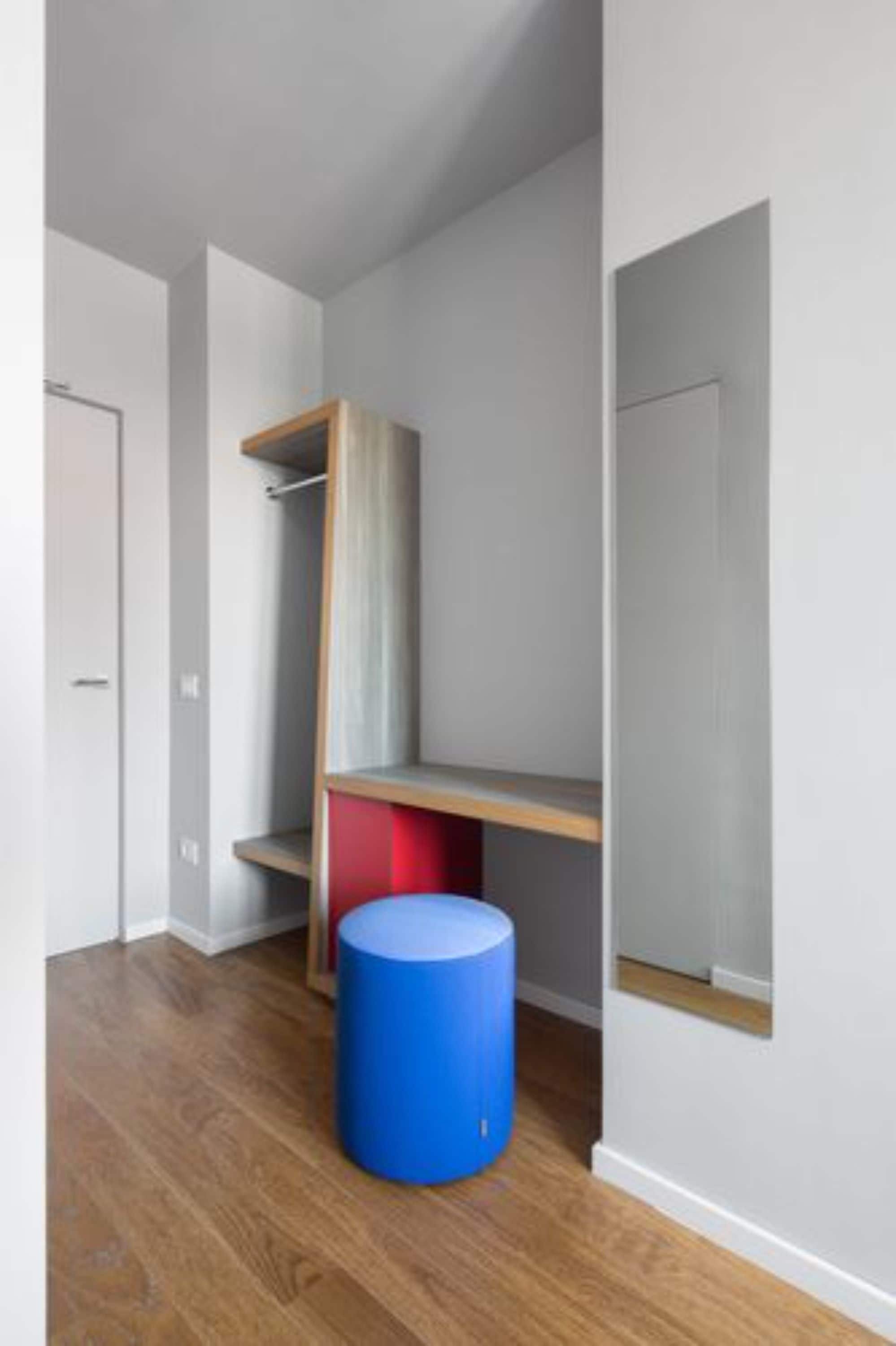 Una stanza con pavimento in legno, un oggetto cilindrico blu, un mobile rosso e grigio e una porta bianca.