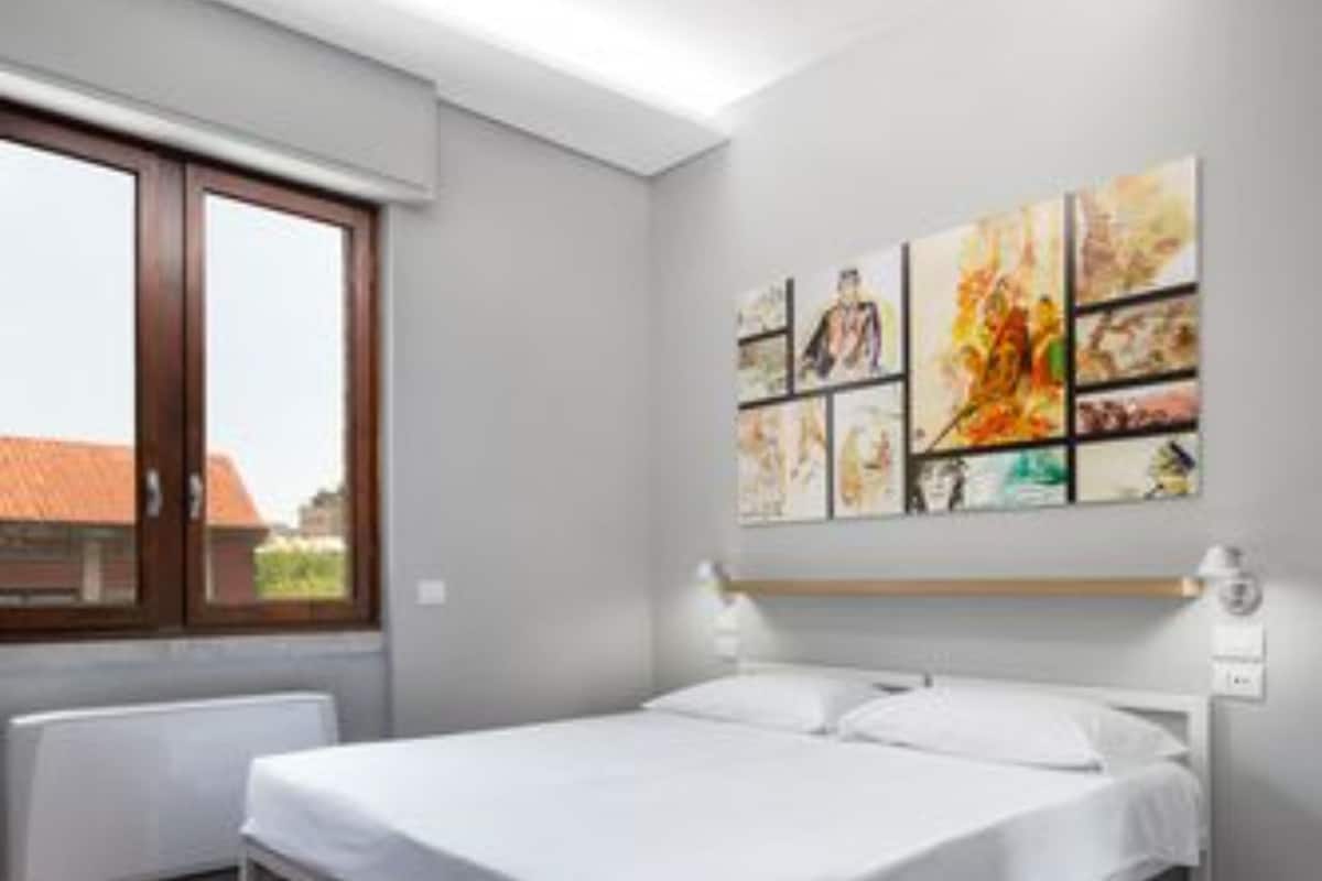 A bedroom with a large bed, a window with a view of buildings, and a wall with a series of artworks.