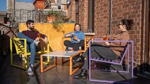 Tres personas sentadas a una mesa, con una pared amarilla y un edificio de ladrillo al fondo.