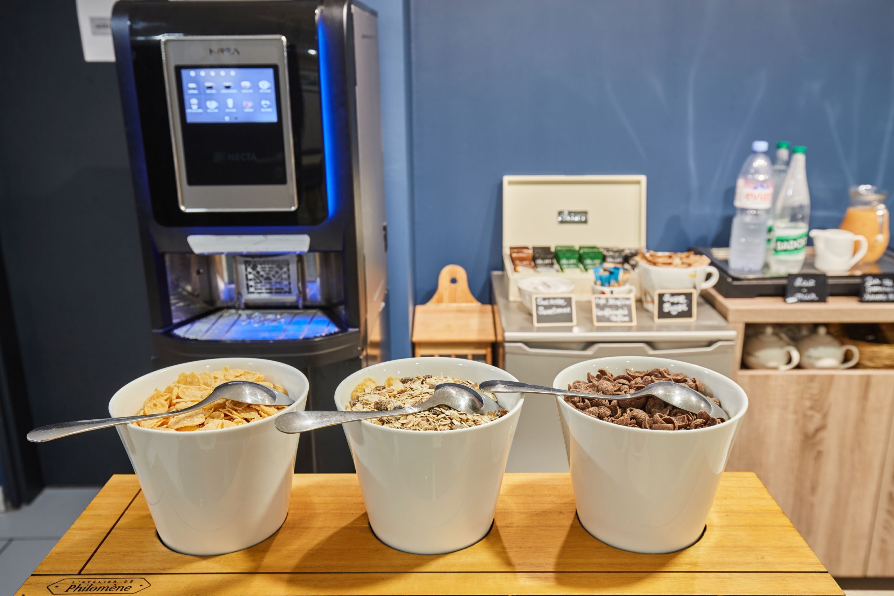 Drei weiße Schüsseln mit Müsli auf einem Holztisch, im Hintergrund eine Kaffeemaschine und eine Kaffeestation.