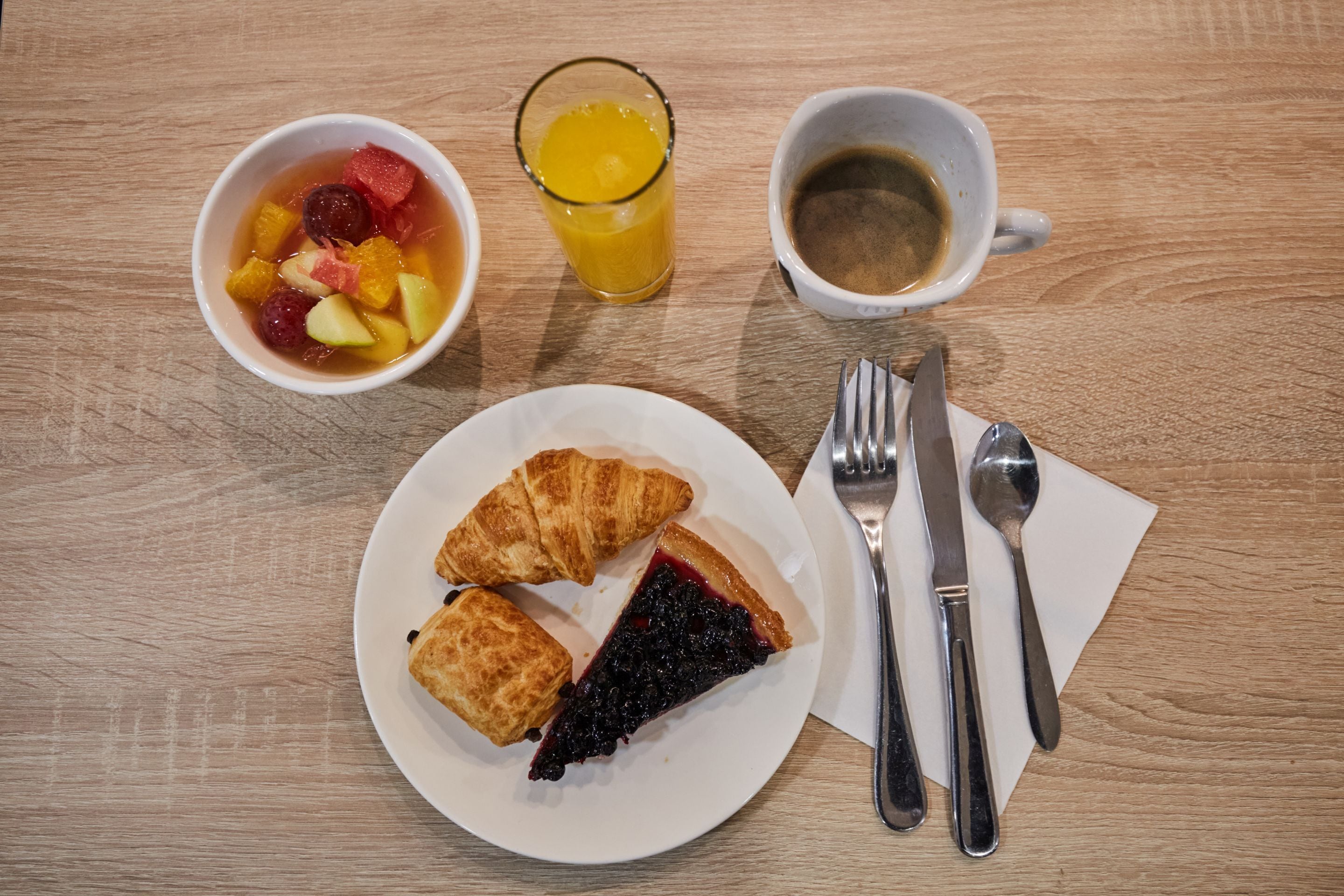 Une table en bois dressée pour le petit-déjeuner, avec un croissant, une tarte aux myrtilles, un saladier de fruits, du jus d’orange et du café.