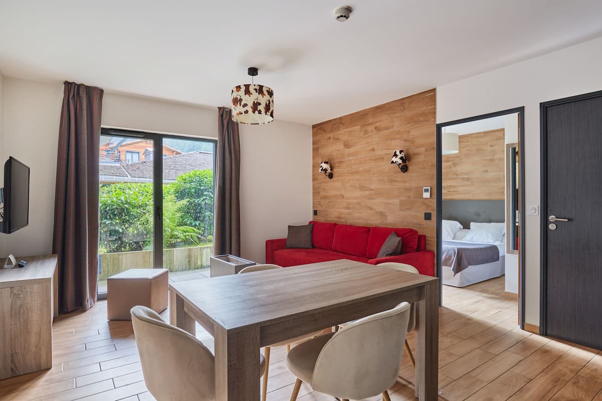 A modern living room with a wooden dining table, a red sofa, and a large window offering a view of greenery outside.
