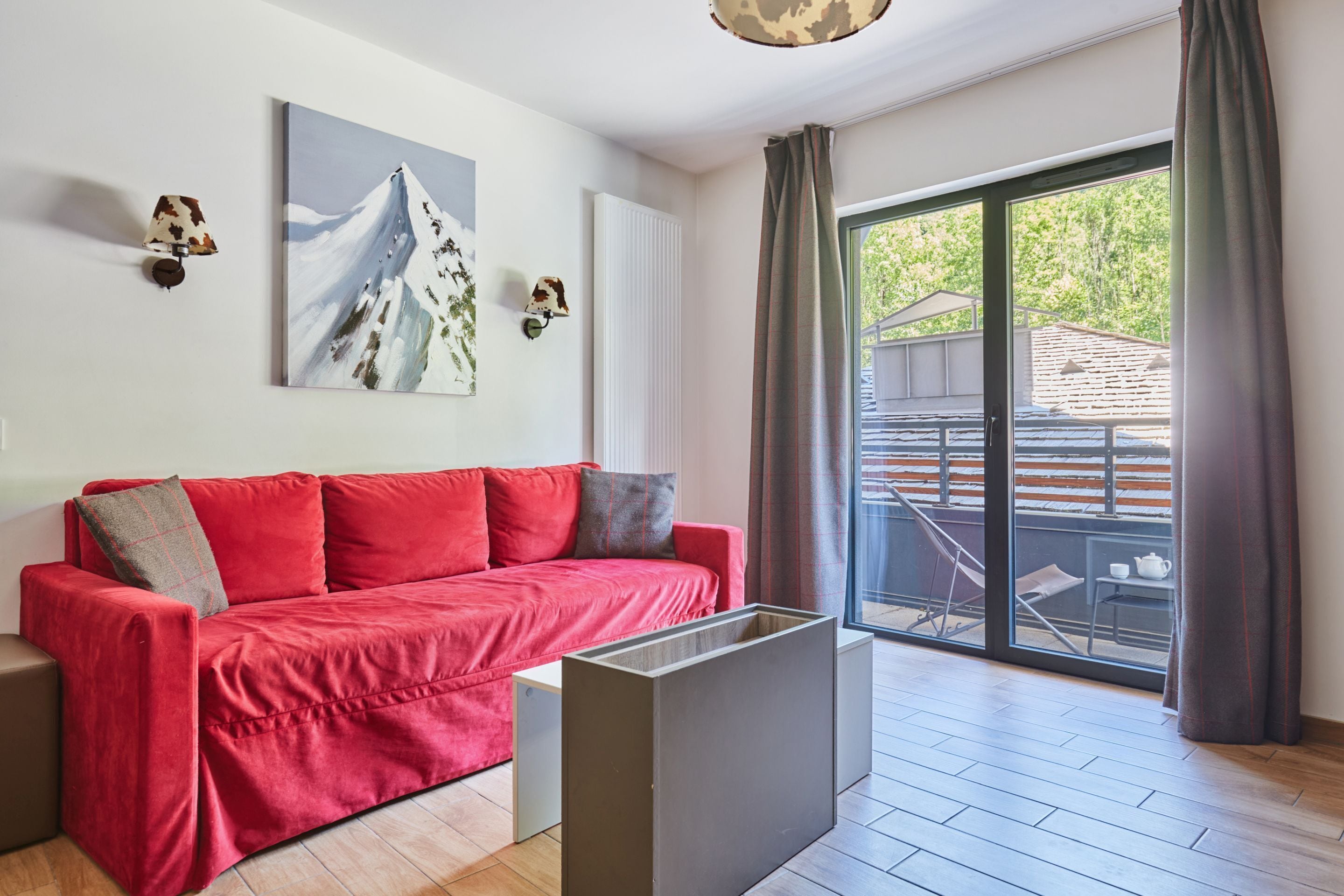 A modern living room with a red sofa, a painting of a mountain, and a view of the outdoors through a glass door.