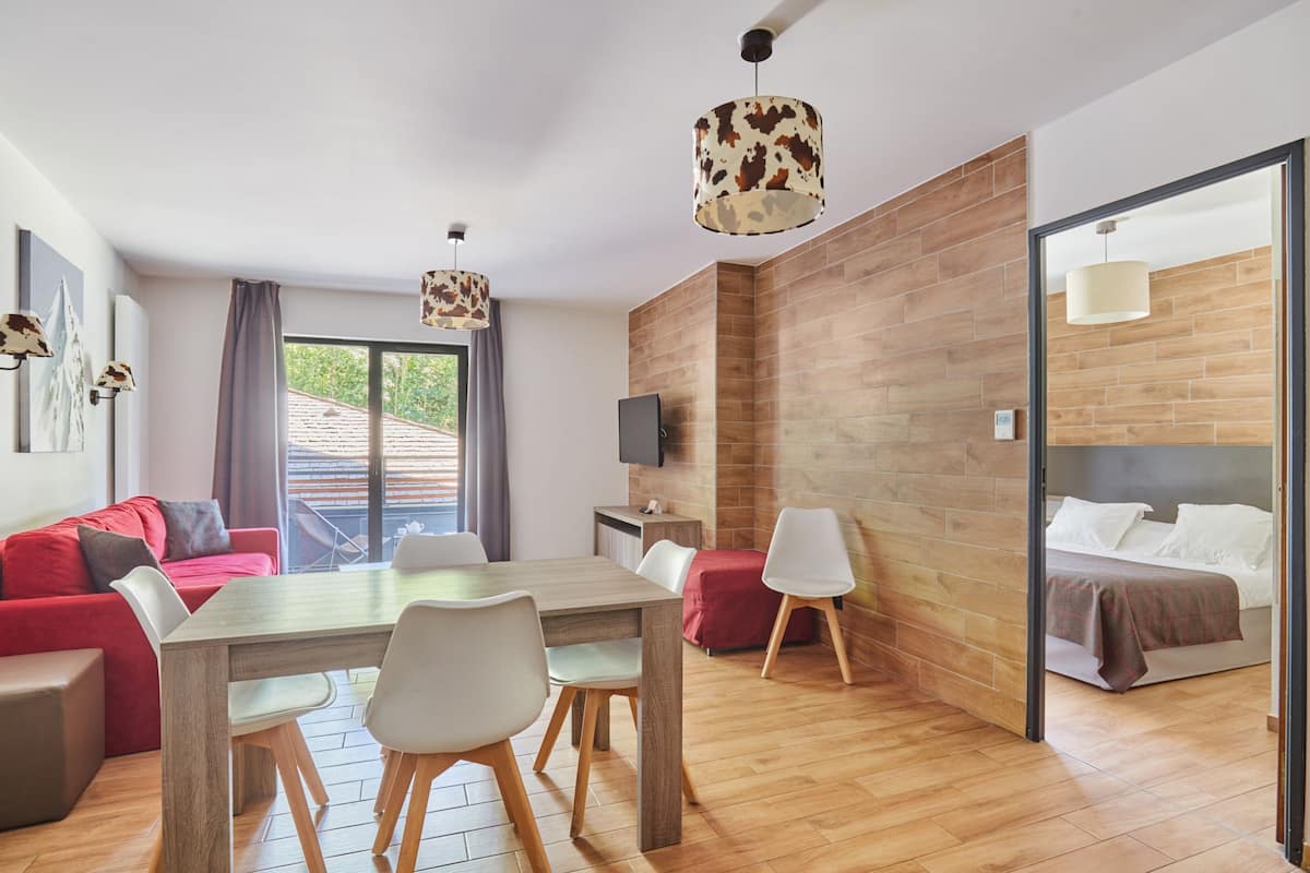 A modern room with a wooden floor, a dining table with chairs, a red sofa, a bed with a white headboard, and a TV mounted on the wall.
