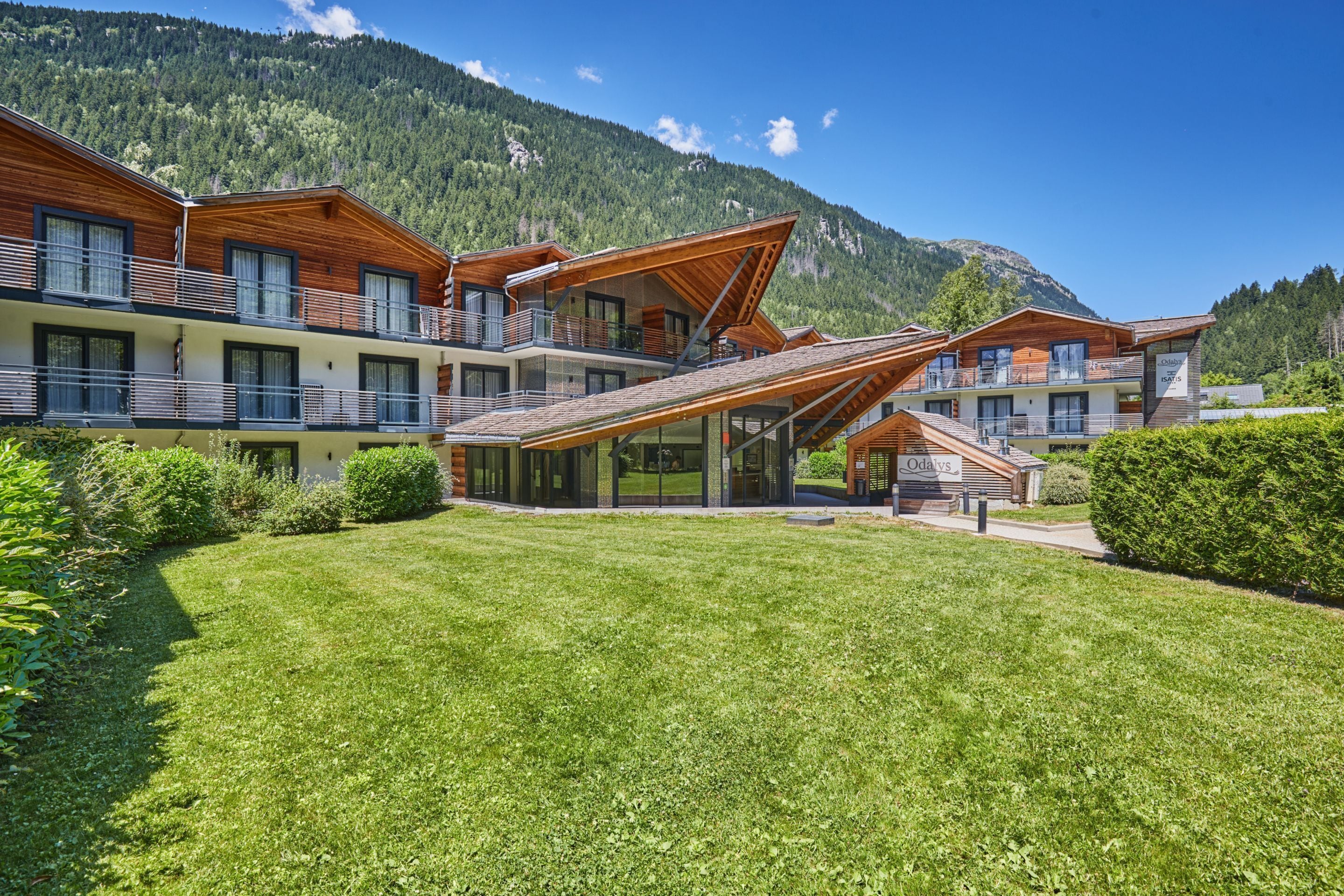 Un hotel moderno con varios balcones de madera, un césped bien cuidado y montañas al fondo.