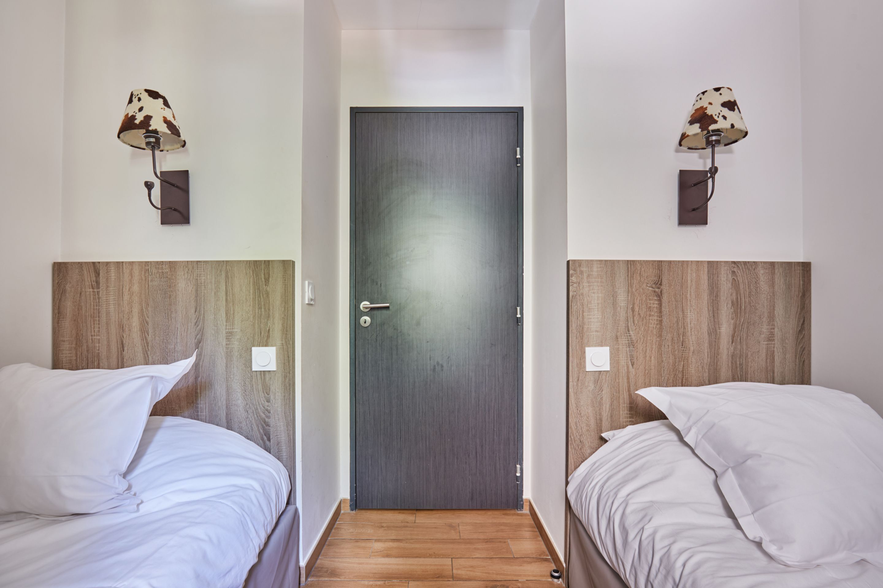 Habitación de hotel con dos camas, cada una con ropa blanca y cabeceros de madera. A ambos lados de una puerta de madera oscura hay lámparas de pared con pantallas de piel de vaca.