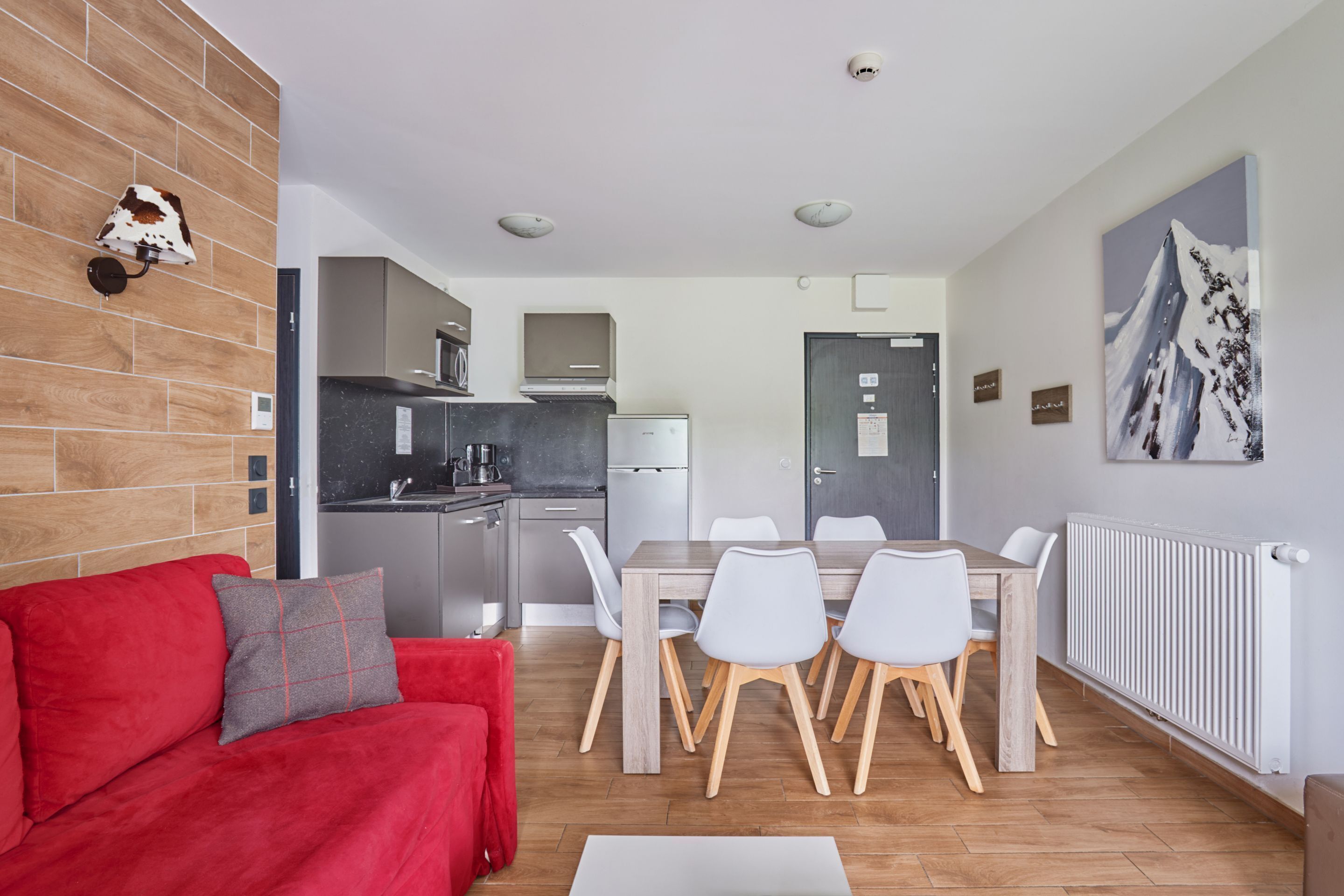 Una cocina moderna con refrigerador, estufa y microondas. Hay un comedor con una mesa y sillas. Se ve un sofá rojo y una almohada gris en primer plano.