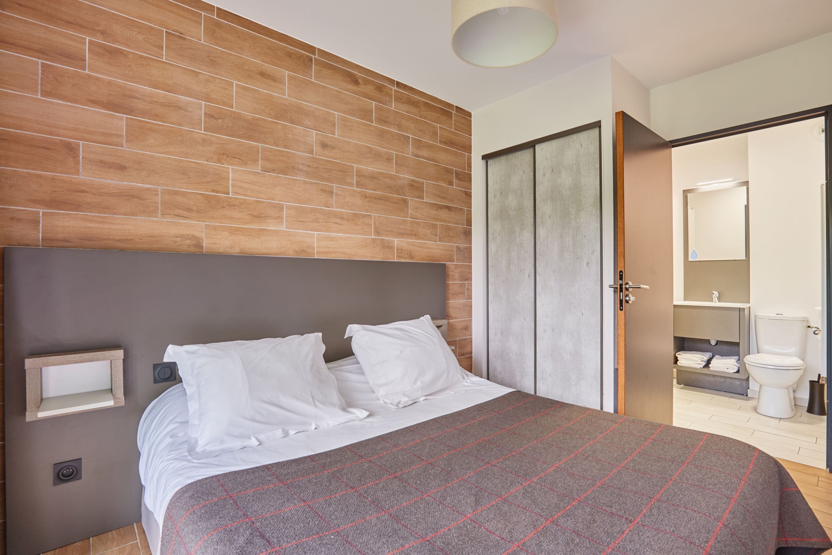 A bedroom with a wooden headboard, a bed with white bedding, a bedside table, and a view into the bathroom.