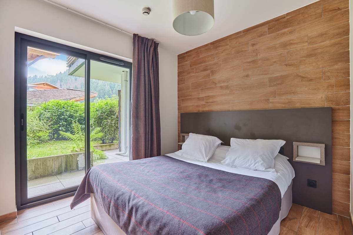 A modern bedroom with a large bed, a wooden accent wall, and a sliding glass door leading to a balcony with a view of greenery and buildings.