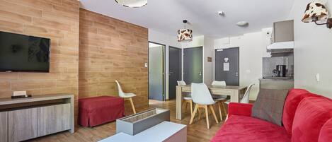 A modern living room with a red sofa, a white coffee table, and a TV mounted on the wall. There is a wooden bench and a dining area with chairs and a table.