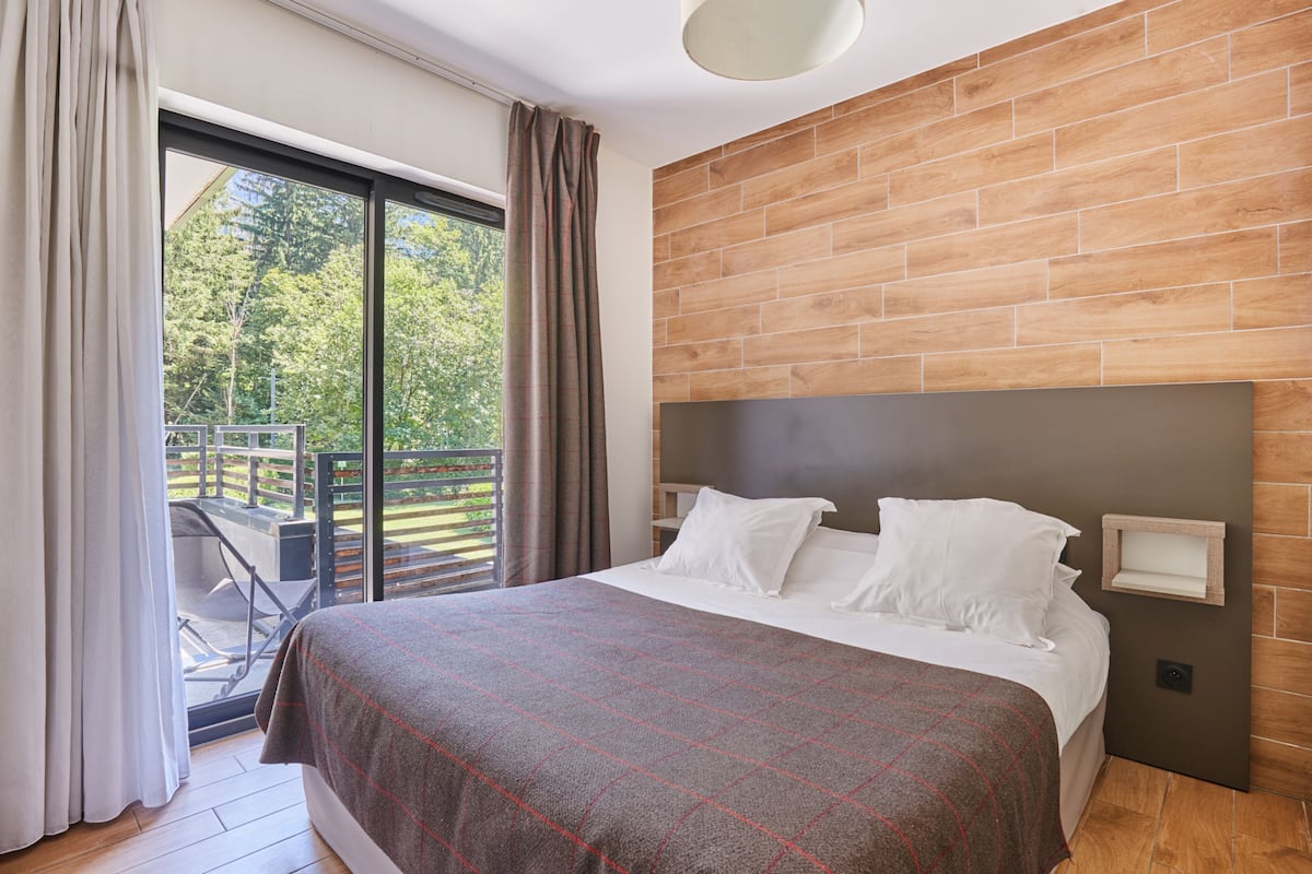 A modern hotel room with a large bed, a wooden headboard, a balcony with a view of trees, and a sliding glass door.