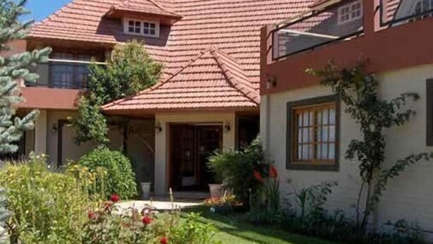 A two-story house with a red-tiled roof, a front porch, and a garden with various plants and flowers. - Amparo Hosteria (Neuquén)