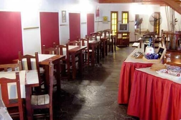 Un área de comedor con mesas y sillas de madera, un mostrador cubierto con mantel rojo y diversos objetos, y un televisor sobre un pedestal.