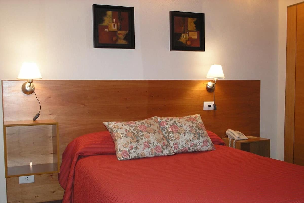 Ein Hotelzimmer mit einem Holzkopfteil, zwei Wandleuchten, einem Telefon und einem blumigen Kissen auf dem Bett.