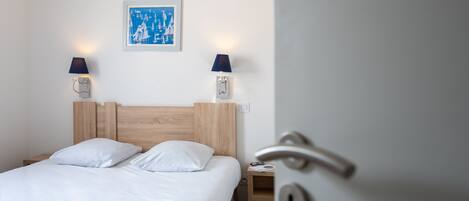 Een hotelkamer met een houten hoofdeinde, witte beddengoed, twee wandlampen, een lijst met een afbeelding en een deur met een handvat.