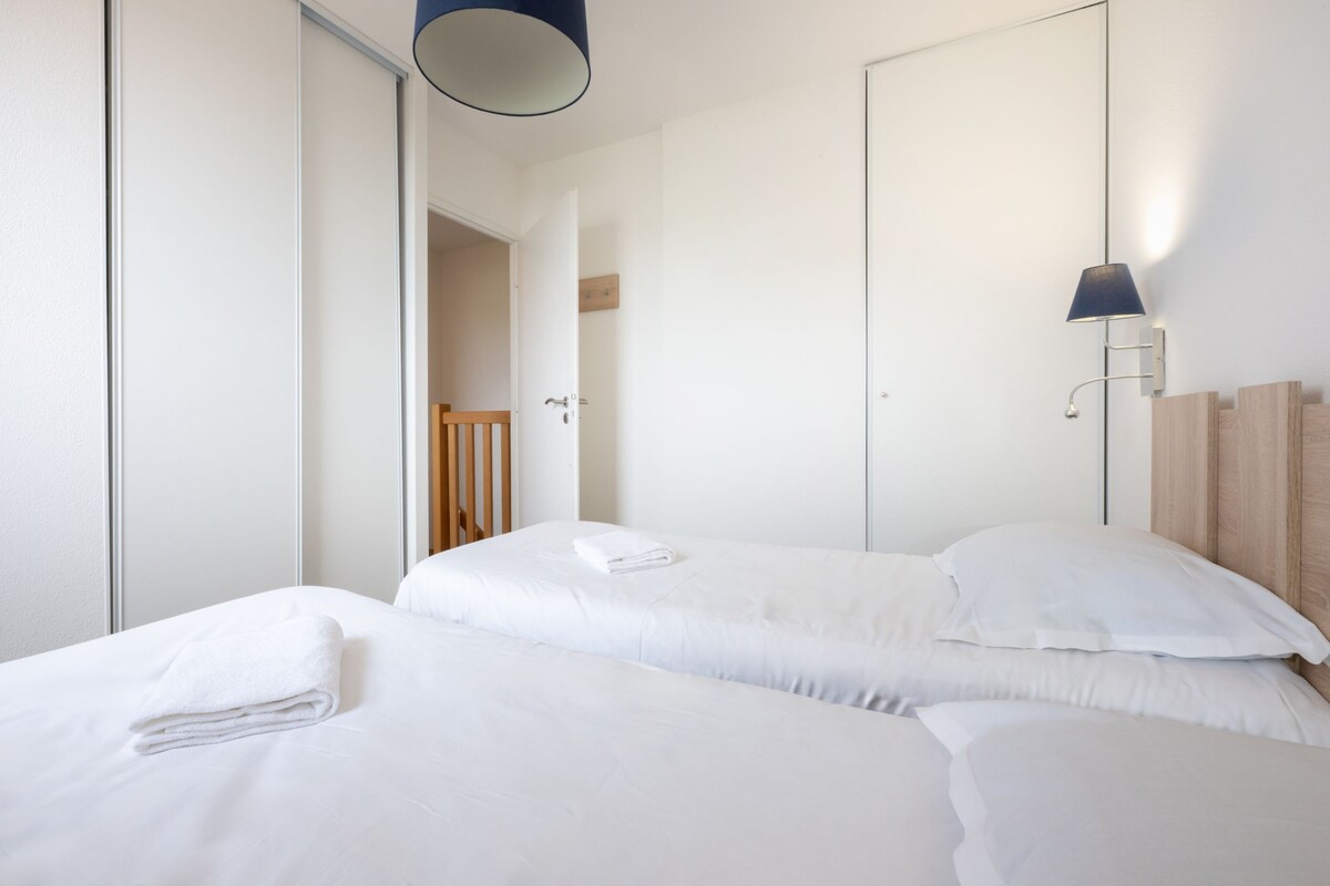 Habitación de hotel con dos camas, cabecero de madera, lámpara de pared y una puerta que da a otra habitación.