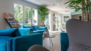 A modern living room with a blue sofa, a grey armchair, and a coffee table. There is a display of books and a large window providing a view of the outdoors. - Résidence Odalys Domaine des Pins - Bois de la Chaise (Noirmoutier-en-l'Ile)