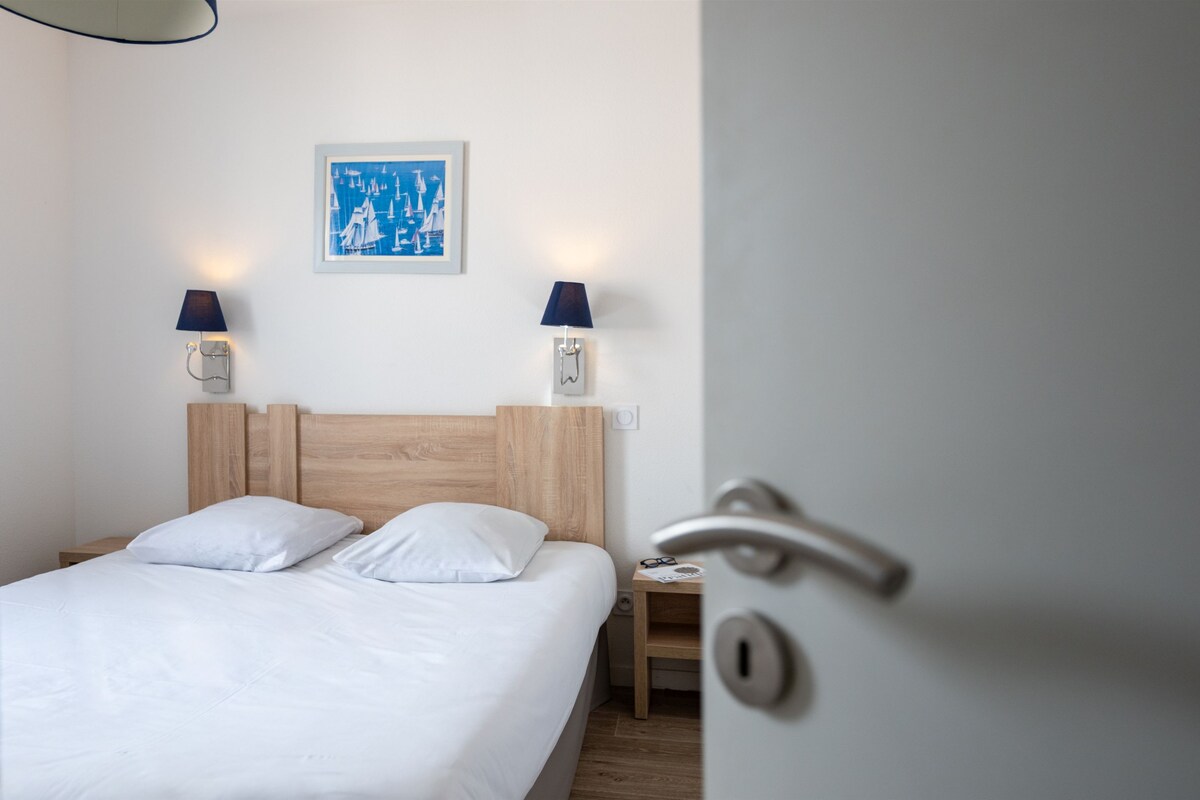 Habitación de hotel con cabecera de madera, ropa de cama blanca, dos lámparas fijadas a la pared, un cuadro enmarcado y una puerta con manija.