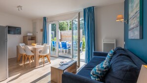 A modern living room with a sofa, a dining table with chairs, and a kitchen area with a TV. - Résidence Odalys Domaine des Pins - Bois de la Chaise (Noirmoutier-en-l'Ile)
