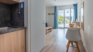 A modern kitchen with a black countertop and wooden cabinets, leading to a living area with a white chair and a table. - Résidence Odalys Domaine des Pins - Bois de la Chaise (Noirmoutier-en-l'Ile)