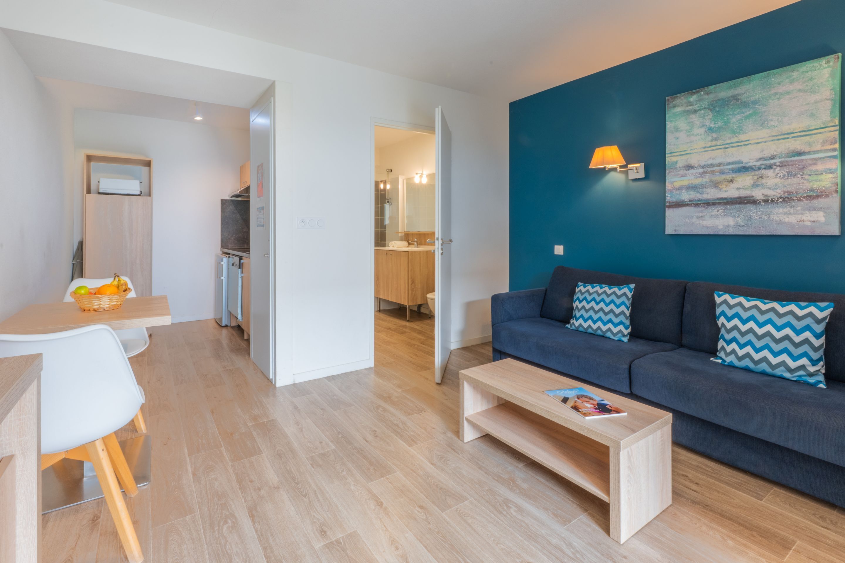 A modern living room with a blue accent wall, a sofa with patterned cushions, a wooden coffee table, and a dining area with a white table and chairs.