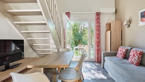 Een moderne woonkamer met een houten eettafel, witte stoelen, een grijze bank met rode kussens, een televisie en een trap.