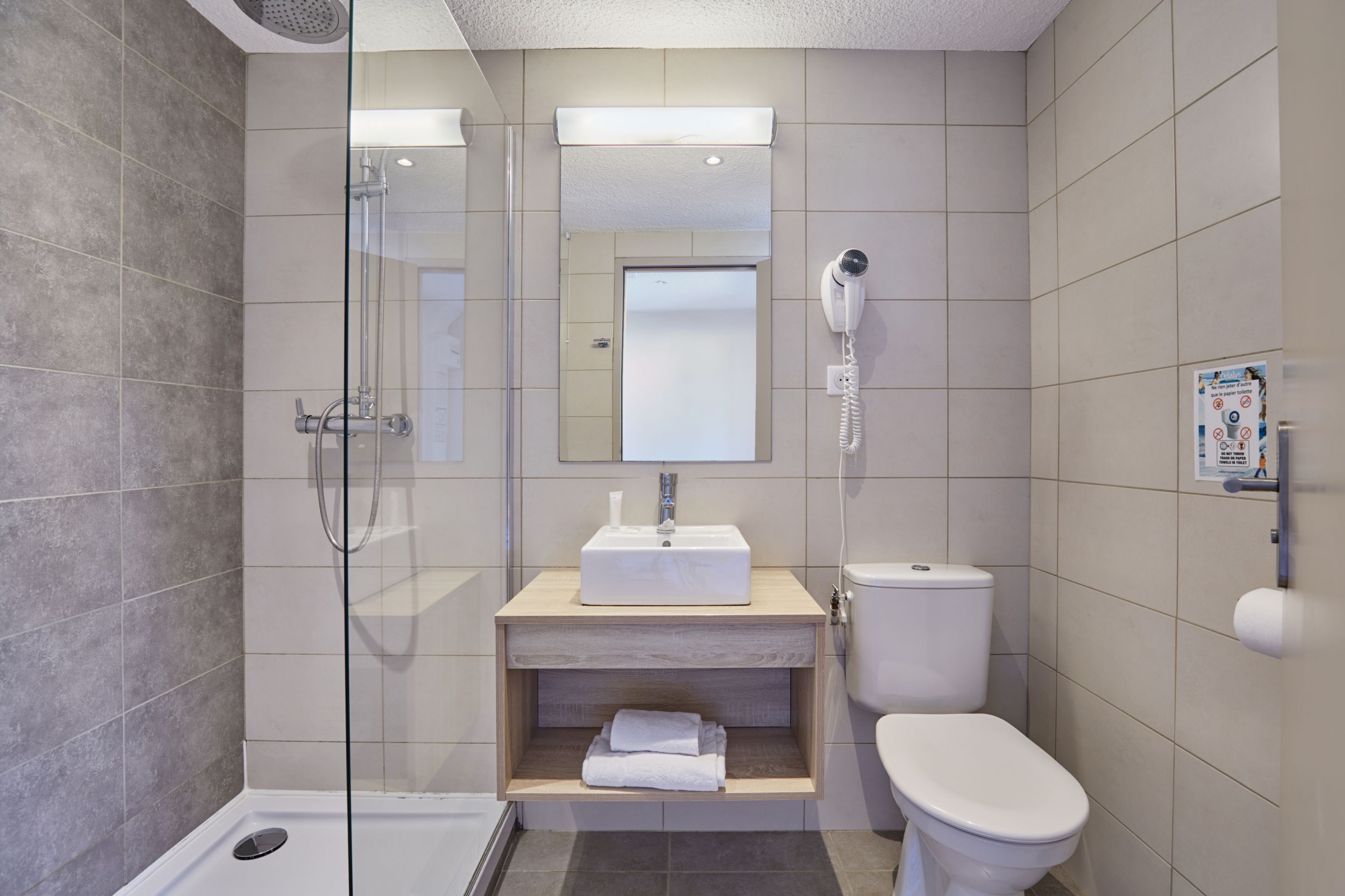 A modern bathroom with a glass shower enclosure, a white sink with a mirror above, a toilet, and a hairdryer mounted on the wall.