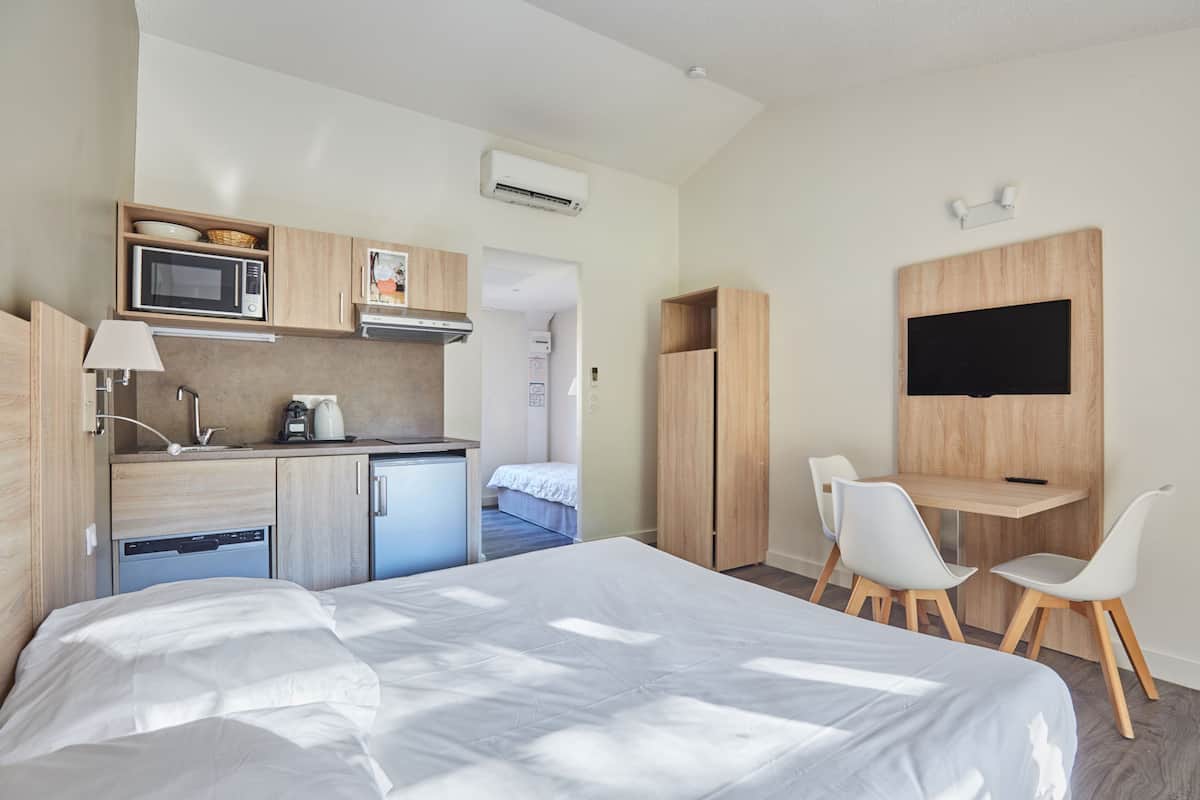 A modern hotel room with a bed, kitchenette, and a desk with chairs.