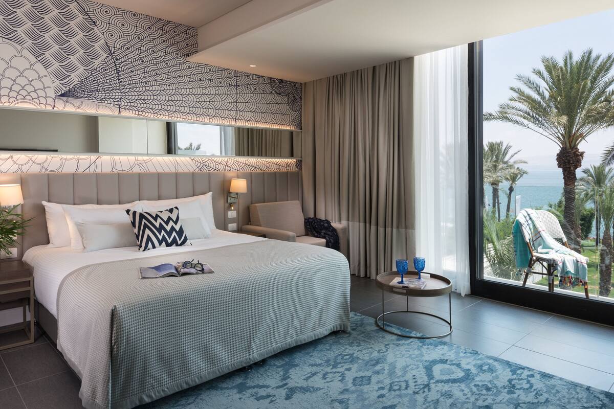 Ein modernes Hotelzimmer mit einem großen Bett, einem Nachttisch, einem kleinen runden Tisch und Blick auf Palmen und das Meer.